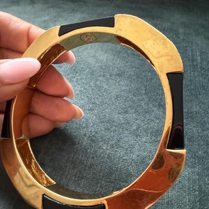 Puede incluir: Una pulsera rígida circular con segmentos alternos de metal dorado pulido y resina negra lisa. El interior de la banda de metal dorado muestra un pequeño grabado de logotipo circular. El diseño presenta un borde geométrico con facetas.