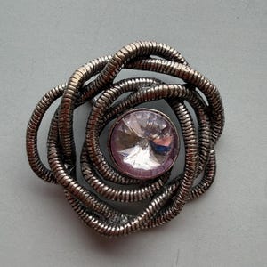 Op de afbeelding: Zilverkleurige broche met een centrale, facetgeslepen, roze edelsteen. Het ontwerp van de broche heeft een geweven, touwachtig patroon dat een cirkelvorm vormt rond de edelsteen. De achtergrond is effen lichtgrijs.