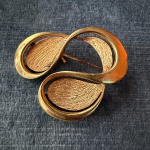 Puede incluir: Broche vintage dorado con diseño abstracto. La broche presenta tres elementos interconectados en forma de lágrima con superficies texturizadas. Un accesorio clásico.
