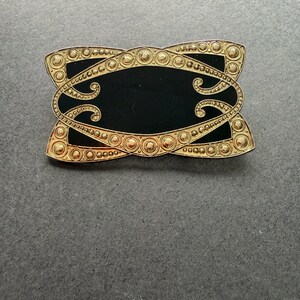 Op de afbeelding: Een rechthoekige goudkleurige broche met een zwart midden. De broche heeft een sierlijk ontwerp met een reeks kleine cirkels en wervelende patronen. De gouden rand heeft een getextureerd uiterlijk, wat diepte aan het ontwerp toevoegt.
