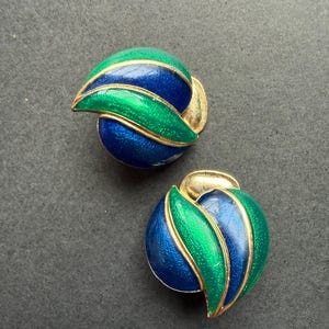 Op de afbeelding: Een paar clip-oorbellen met een afgerond, gelaagd ontwerp met afwisselende secties van levendig blauw en heldergroen emaille. De gebogen segmenten zijn omlijnd met goudkleurig metaal, wat zorgt voor een gladde en gepolijste afwerking.
