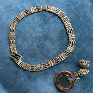 May include: A gold-toned necklace with a rectangular link design, a matching brooch with a green center, and a pair of earrings with pearl accents. The necklace has a tag that reads "98.00 DM". The jewelry is displayed on a blue surface.