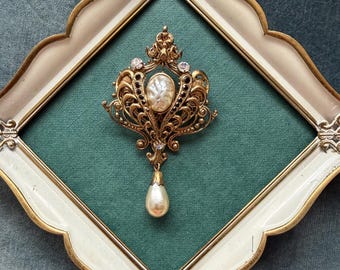 Könnte beinhalten: Eine verzierte, goldfarbene Brosche mit einer zentralen ovalen Perle und einem herabhängenden Perltropfen, vor einem türkisgrünen Hintergrund in einem weiß-goldfarbenen Rahmen. Die Brosche hat aufwendige Filigranarbeiten und funkelnde Akzente.