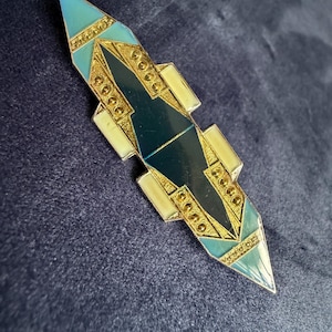 Op de afbeelding: Een lange, puntige broche met een geometrisch ontwerp, gouden metalen accenten, donkergroene emaille panelen en lichtblauwe en crèmekleurige emaille secties. Het sieraad bevat gestructureerde gouden metalen details met kleine cirkelvormige patronen.
