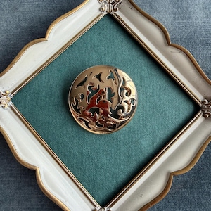 Op de afbeelding: Een goudkleurige, ronde broche met een ingewikkeld uitgesneden ontwerp met een centrale figuur en omringend gebladerte. De broche is geplaatst tegen een teal achtergrond in een wit en goudkleurig sierlijk frame.