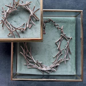 Könnte beinhalten: Ein silberfarbenes Armband und eine passende Halskette, beide aus miteinander verbundenen, schlanken, stabartigen Segmenten gefertigt, die in einem chaotischen, zweigartigen Muster angeordnet sind. Jedes Stück hat kleine, dezente blaue Akzente.