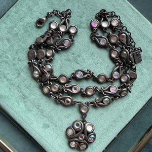Op de afbeelding: Een meerrijige ketting met donkergrijze metalen schakels in krulvormige motieven. De ketting is versierd met talloze ronde, iriserende en heldere cabochonstenen en eindigt in een centrale hanger in de vorm van een bloem.