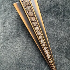 May include: A bronze-toned metal brooch with a geometric design. The brooch features a central row of clear, round rhinestones. Two bronze-coloured metal wings extend from the top. The rhinestones decrease in size towards the pointed end.