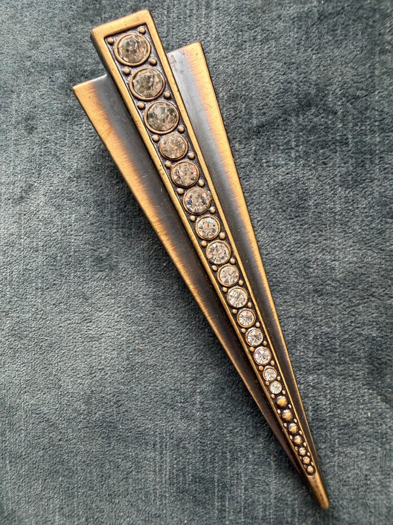May include: A bronze-toned metal brooch with a geometric design. The brooch features a central row of clear, round rhinestones. Two bronze-coloured metal wings extend from the top. The rhinestones decrease in size towards the pointed end.
