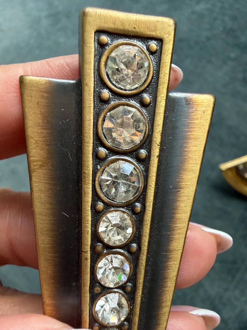 May include: Art Deco style metal hardware with a bronze finish. The hardware features a vertical row of five clear, round, faceted crystal accents. The edges are framed with a gold-tone metal.