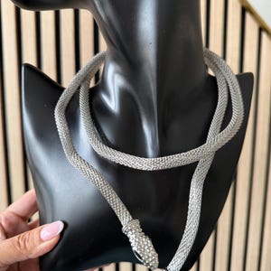Op de afbeelding: Zilverkleurige slangenketting, gepresenteerd op een zwarte mannequinhoofd. De ketting is gemaakt van een mesh-achtig materiaal en heeft een slangenkop- en staartsluiting. De slangenkop is gedetailleerd met kleine, sprankelende versieringen.