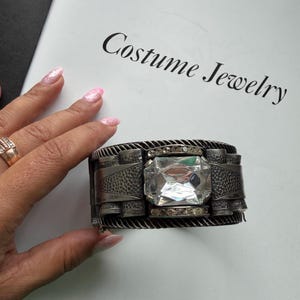 Puede incluir: Un brazalete ancho de metal en tono oscuro con una gran piedra central rectangular de facetas transparentes, flanqueada por filas de cristales redondos más pequeños. La superficie metálica presenta un acabado texturizado y martillado.