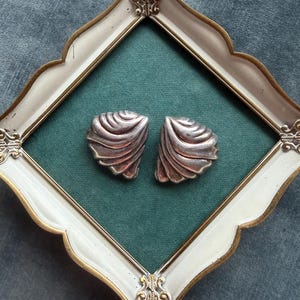 May include: A pair of silver-toned earrings with a fan-like, ribbed shell design. The earrings feature a curved, layered structure with a metallic finish, displayed against a dark green background within a decorative, ornate cream and gold frame.