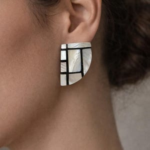 Op de afbeelding: Close-up van een wit en zwart geometrische oorbel. De oorbel heeft een halve cirkelvorm met een rasterachtig ontwerp. De witte delen hebben een parelmoerglans. De zwarte lijnen creëren een moderne, artistieke uitstraling.
