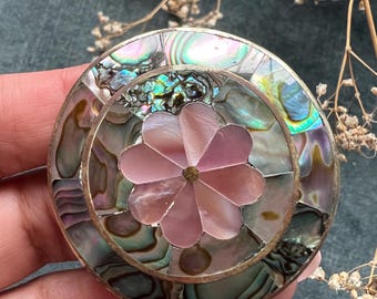 Könnte beinhalten: Runde, dekorative Brosche mit silberfarbenem Metallrahmen. Die Brosche zeigt ein zentrales Blumendesign in Rosa, umgeben von schillernden Abalone-Muschelteilen in Grün-, Blau- und Lilatönen. Die Brosche wird von einer Hand gehalten.