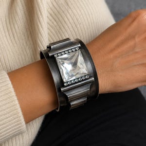 May include: A wide cuff bracelet made of dark, brushed gunmetal-toned metal. The center features a large, square-cut clear crystal stone, flanked by two horizontal rows of small, sparkling clear rhinestones set into the metal frame.