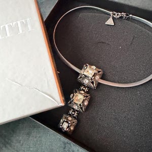 May include: A silver necklace with three square pendants, each with a central light-coloured stone and surrounding smaller stones. The necklace is in a black box next to a white box with the letters "TTI" on it.