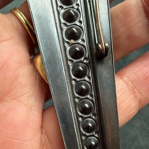 May include: Close-up of a silver brooch with a decorative border of dark, rounded studs. The brooch has a pin closure on the back and a gold-coloured ring is visible in the background. The brooch is likely vintage or antique.