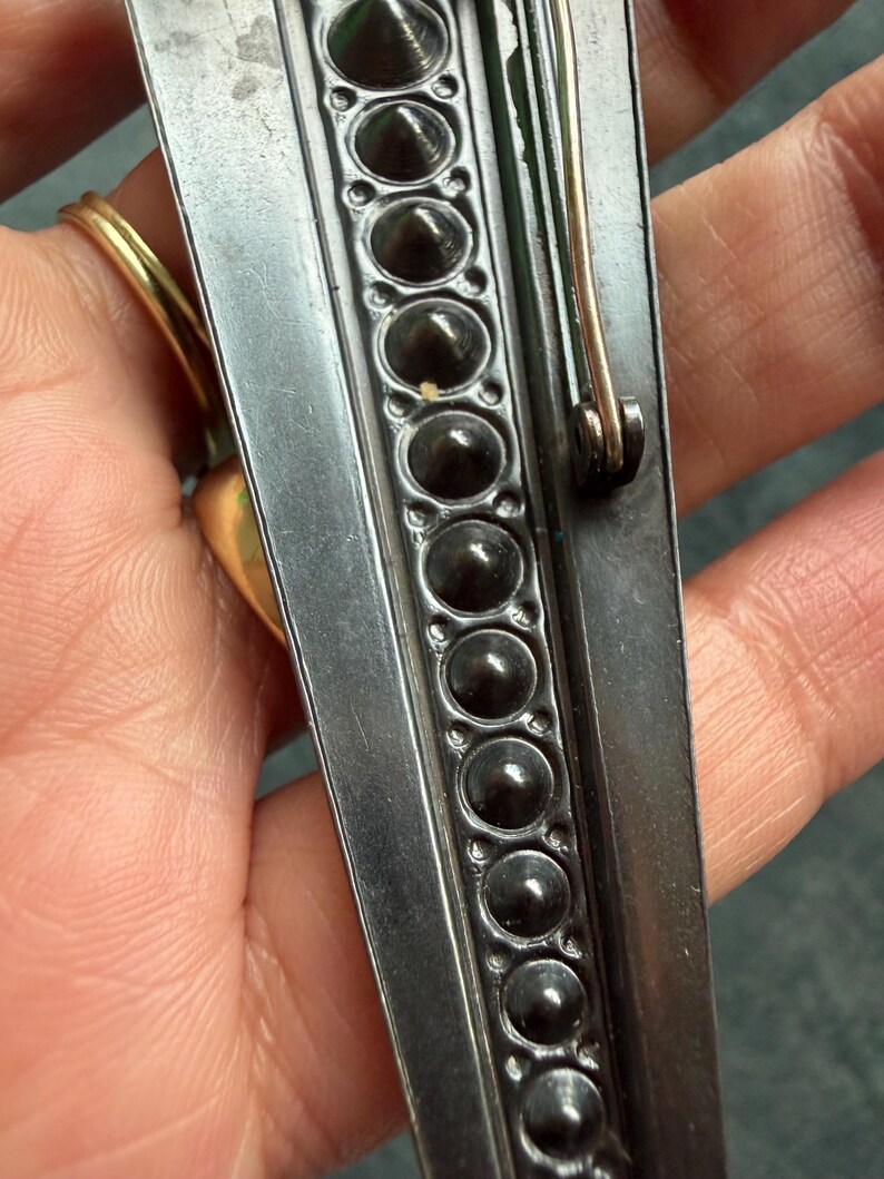 May include: Close-up of a silver brooch with a decorative border of dark, rounded studs. The brooch has a pin closure on the back and a gold-coloured ring is visible in the background. The brooch is likely vintage or antique.