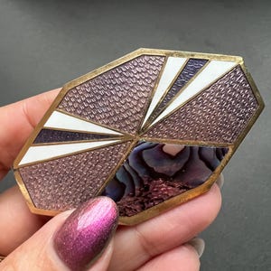 Op de afbeelding: Een geometrische broche met een paars, wit en gouden ontwerp. De broche is gemaakt van metaal en heeft een getextureerd oppervlak. Het midden van de broche heeft een stukje iriserende abalone-schelp.