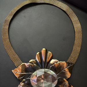 Op de afbeelding: Een gouden ketting met een groot, facetgeslepen kristal in het midden, omgeven door een bloemmotief. Het bloemmotief is van metaal en heeft een donkere, verweerde afwerking.