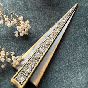 May include: A vintage-style, triangular hair clip with a gold-tone metal frame. The clip features a central row of clear, round rhinestones and a contrasting silver-tone finish on the outer edges. The clip is set against a dark teal background.