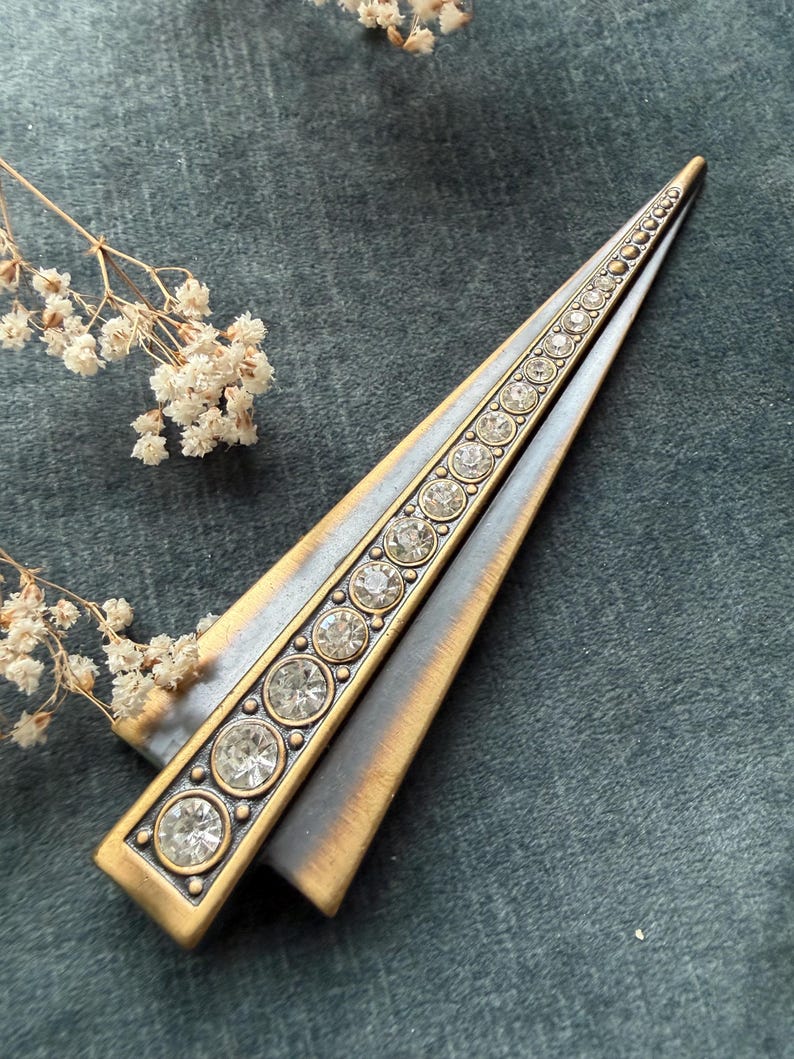 May include: A vintage-style, triangular hair clip with a gold-tone metal frame. The clip features a central row of clear, round rhinestones and a contrasting silver-tone finish on the outer edges. The clip is set against a dark teal background.