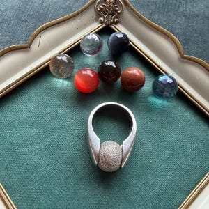 Può includere: Un anello d'argento con un centro sferico strutturato è esposto con una collezione di perline di pietre preziose colorate. Le perline includono tonalità chiare, rosse, marroni e blu. Gli oggetti sono disposti su una superficie verde all'interno di una cornice decorativa.