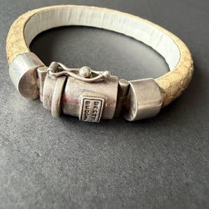 Puede incluir: Una pulsera beige texturizada con un patrón similar a la piel de serpiente y un forro interior blanco. La pieza cuenta con un mecanismo de cierre de metal plateado con una placa rectangular grabada con el texto BESTTO BUDDHA, asegurado por un pestillo.