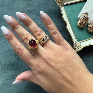 Op de afbeelding: Een hand met drie sieraden: een gouden ring met een grote, ovale, rode edelsteen, een gouden schakelring en een paar gouden oorbellen met kleine, kleurrijke edelstenen. De hand heeft lange, verzorgde nagels met witte punten.