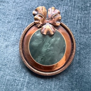 Op de afbeelding: Een vintage broche met een koperkleurig frame en een donkergroen midden. De broche heeft een decoratieve strik aan de bovenkant en een getextureerde, cirkelvormige rand. Het ontwerp is sierlijk en gedetailleerd.