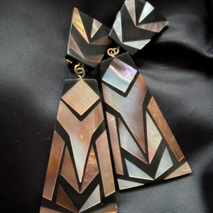 Op de afbeelding: Geometrische oorbellen met iriserende schelpinleg. De oorbellen hebben een zwarte achtergrond met een mozaïek van schelpstukken in bruin-, zilver- en wittinten. De oorbellen hebben goudkleurige haken.