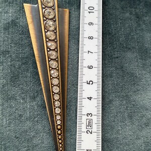 May include: Art Deco style metal brooch with a bronze and black finish. The brooch is approximately 10 cm long and features a row of clear, round rhinestones down the centre. The design is a stylized arrow or fan shape.