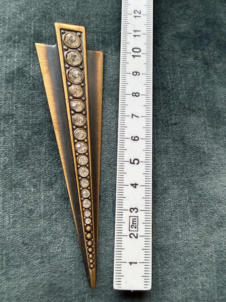 May include: Art Deco style metal brooch with a bronze and black finish. The brooch is approximately 10 cm long and features a row of clear, round rhinestones down the centre. The design is a stylized arrow or fan shape.