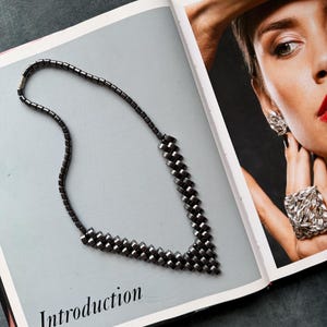 Op de afbeelding: Een donkergrijze ketting met een geometrisch ontwerp, liggend op een pagina van een boek met het woord "Introduction". De ketting heeft een V-vormige hanger. Het gezicht van een vrouw met sieraden is zichtbaar aan de rechterkant van het open boek.
