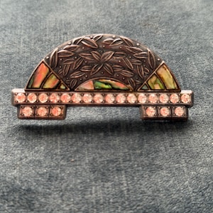 Op de afbeelding: Zilverkleurige broche met een halve cirkel ontwerp. Het bovenste gedeelte heeft een bladerenpatroon en iriserende abalone schelp accenten. De onderkant heeft een rij heldere, ronde strass steentjes. Een decoratief sieraad.