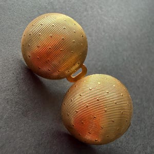 Op de afbeelding: Twee goudkleurige, bolvormige knopen met een getextureerd, geribbeld oppervlak en kleine gestippelde details. De knopen zijn verbonden door een kleine lus. De achtergrond is donkergrijs.
