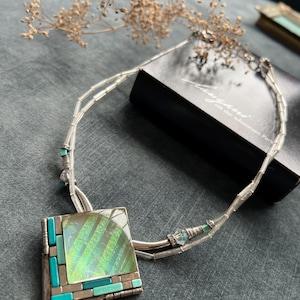 Op de afbeelding: Zilveren ketting met een vierkante hanger met een glinsterend, iriserend midden en turquoise en zilveren accenten. De ketting heeft een delicate kralenketting en wordt gepresenteerd op een grijze ondergrond.