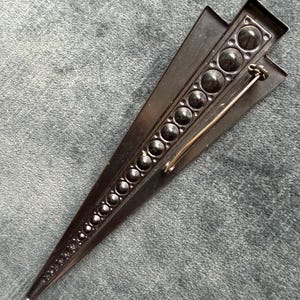 May include: A dark, geometric brooch with a triangular design. The brooch has a series of graduated, circular embellishments down the centre and a pin closure. The brooch is a dark colour, possibly black.