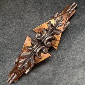 Op de afbeelding: Art Deco-stijl broche met een centraal zilverkleurig bladerontwerp. De broche heeft een geometrische vorm met brons- en goudtinten en is versierd met kleine, sprankelende strass-steentjes langs de randen.