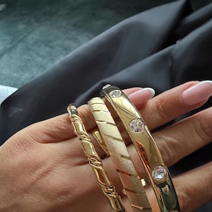 Op de afbeelding: Vier gouden armbanden in verschillende ontwerpen. Eén is gedraaid, een andere heeft een textuurpatroon en de derde heeft drie heldere edelstenen. De armbanden worden op een hand getoond en tonen hun verschillende stijlen en afwerkingen.