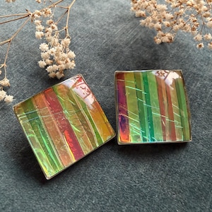 Op de afbeelding: Twee vierkante oorbellen met iriserende, kleurrijke, verticale strepen in groen, rood en geel. De oorbellen hebben een zilverkleurige metalen rand. De oorbellen worden gepresenteerd op een donkergrijze ondergrond.