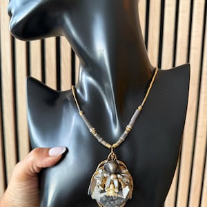 Op de afbeelding: Een goudkleurige ketting met een grote hanger. De hanger heeft een centrale grijze steen, omringd door gouden en zilveren details. De ketting wordt gepresenteerd op een zwarte mannequin. De ketting is van kralen.