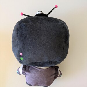 Op de afbeelding: Een knuffel met een grote, vierkante, donkergrijze kop met twee zwarte antennes met kleine roze bolletjes. De kop heeft een klein roze cirkeltje en een groen cirkeltje aan de zijkant. Het lichaam is gedempt paars met donkergrijze accenten.