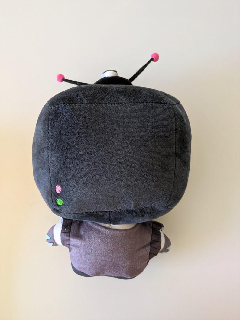 Op de afbeelding: Een knuffel met een grote, vierkante, donkergrijze kop met twee zwarte antennes met kleine roze bolletjes. De kop heeft een klein roze cirkeltje en een groen cirkeltje aan de zijkant. Het lichaam is gedempt paars met donkergrijze accenten.