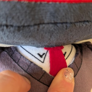 Op de afbeelding: Een knuffel met een donkergrijze, getextureerde kop en een wit kraaggedeelte met een felrode verticale streep. Het kraaggedeelte bevat zwarte stikdetails en een donkergrijs, trui-achtig kledingstuk met zichtbare stiksels langs de halslijn.