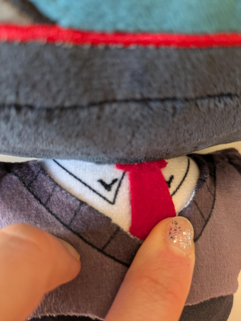 Op de afbeelding: Een knuffel met een donkergrijze, getextureerde kop en een wit kraaggedeelte met een felrode verticale streep. Het kraaggedeelte bevat zwarte stikdetails en een donkergrijs, trui-achtig kledingstuk met zichtbare stiksels langs de halslijn.