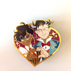 Può includere: Una spilla a forma di cuore in smalto con due personaggi dei cartoni animati che formano un cuore con le mani. La spilla ha un bordo dorato, uno sfondo viola glitterato e accenti di stelle. I personaggi sono vestiti in abiti formali.