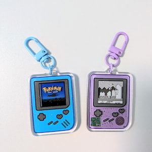 Op de afbeelding: Twee rechthoekige acryl sleutelhangers in de vorm van draagbare videogameconsoles. De ene is lichtblauw met een scherm dat een Pokémon-logo weergeeft, en de andere is paars met een scherm dat een zwart-wit gepixelde spelscène toont.