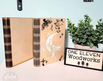 May include: Two wooden bookends with a laser-cut design featuring a crescent moon, stars, a moth, and a hand holding a pendulum. The bookends are made of light wood with black laser-cut details. The text "ONE ELEVEN WOODWORKS" is visible on a separate wooden sign.